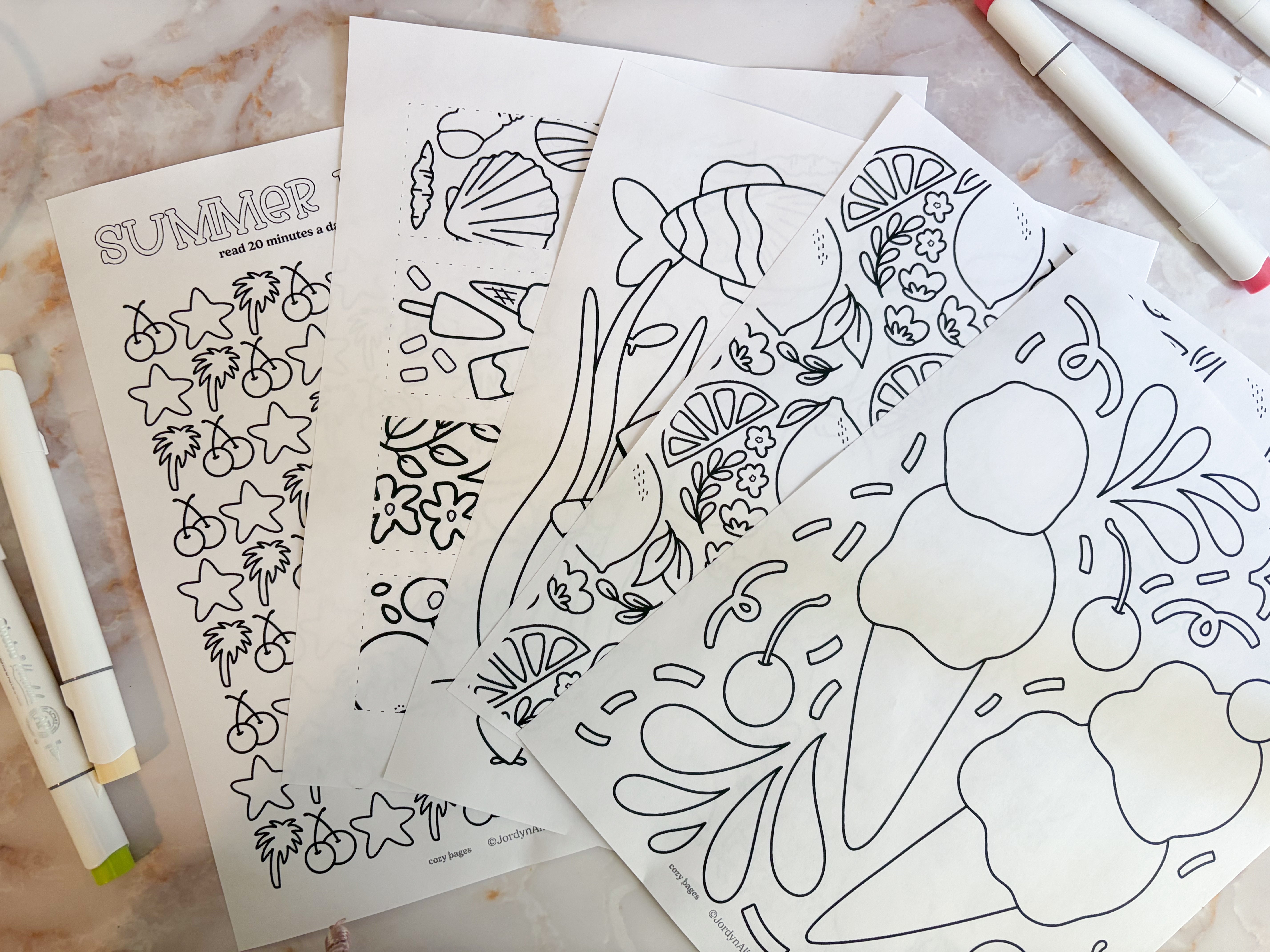 Coloring sheets with summer-themed designs on a marble surface with markers.