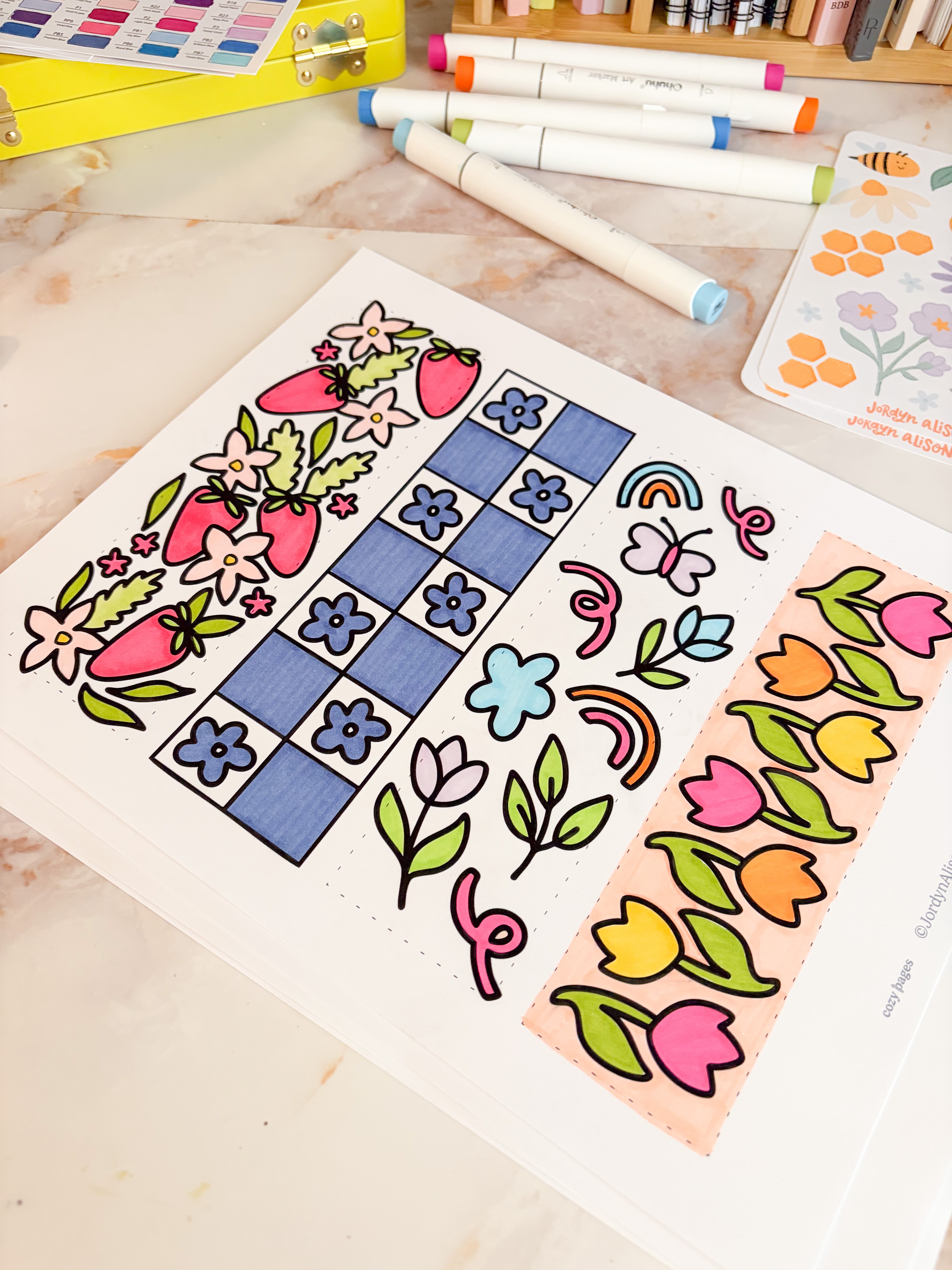 Colorful floral and geometric designs on a sheet of paper with markers and coloring books in the background.